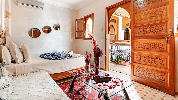 A room with a bed, a glass coffee table with rose petals, a vase with red flowers, and a wooden door with intricate carvings.