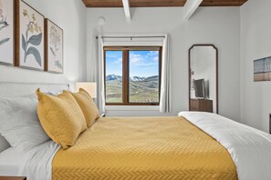 Een slaapkamer met een bed, een raam met uitzicht op bergen, een spiegel en lijstwerken aan de muur.