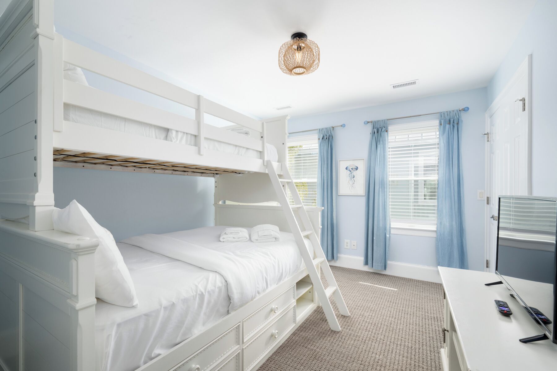 A bunk bed room with a desk and a ladder, a ceiling light, and a window with curtains.