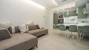 Una sala moderna con sofá y sillones, un comedor con mesa y sillas, y una cocina con electrodomésticos.
