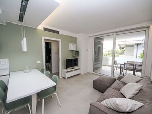 Ein modernes Wohnzimmer mit einem weißen Tisch, grünen Sesseln, einem grauen Sofa, einem Fernseher und einer Glasschiebetür, die zu einem Außenbereich führt.