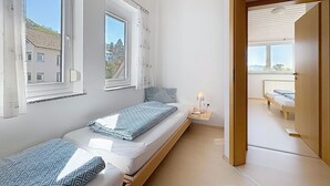 Ein kleines Zimmer mit einem Einzelbett, einem Fenster, einem Nachttisch mit Lampe und Blick nach draußen.