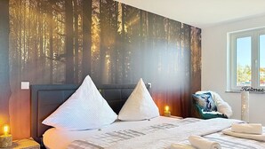 Hotelkamer met een bed, kussens, een nachtkastje met lamp, een raam en uitzicht op bomen.
