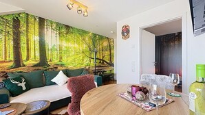 Een woonkamer met een bosdecoratie, een ronde houten tafel met drankjes, een groene bank en een klein bijzettafeltje.