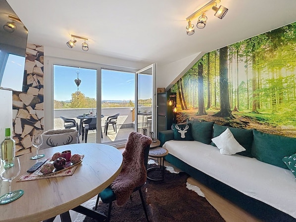 Ein modernes Wohnzimmer mit großem Fenster, einer Essecke mit Tisch für zwei Personen und einer gemütlichen Sitzecke mit Wandgemälde zum Thema Wald.