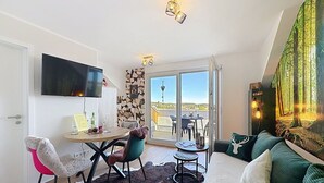 Een moderne woonkamer met een flatscreen-tv, een eethoek met tafel en stoelen, een bank met groene kussens en een groot raam met uitzicht op een bos.