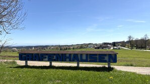 Naambord met 'GRAFENHAUSEN' op een bank in een grasveld, heldere lucht.