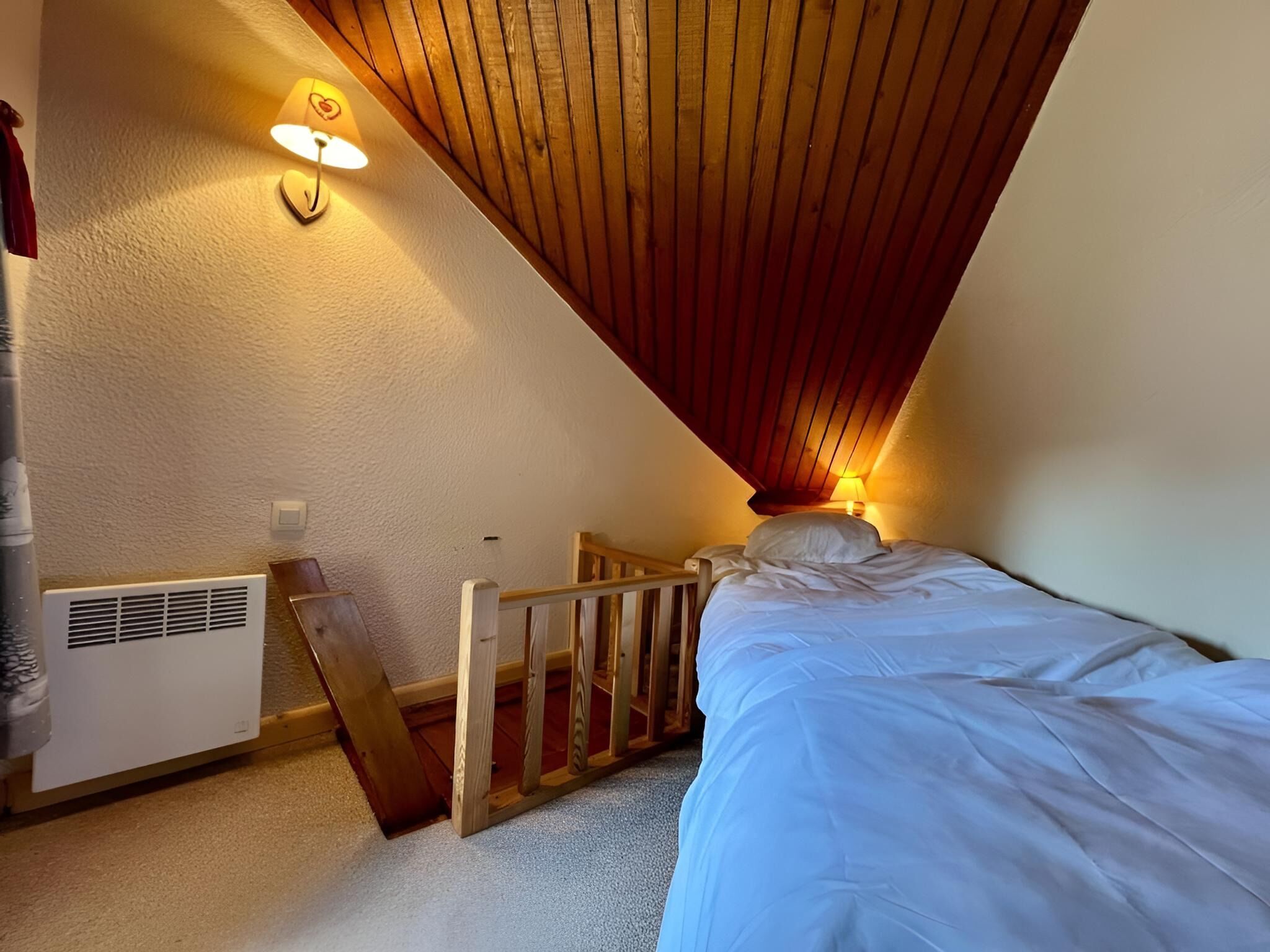 A bedroom with a sloped ceiling, a bed, a nightstand, a wall-mounted lamp, and a radiator.