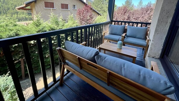 Un balcon avec un banc en bois et deux fauteuils rembourrés, donnant sur une maison et de la verdure.