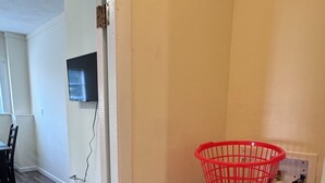 Ett tvättrum med en röd korg på en torktumlare, en dörr som leder till ett vardagsrum med en tv och ett trägolv.