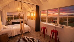 Un dormitorio con una cama grande, una silla colgante, vista al atardecer y una ventana con panorama del paisaje.
