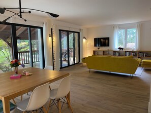 A modern living room with a yellow sofa, a dining table with chairs, a TV mounted on the wall, and a sliding glass door leading to an outdoor area. - Spacious Landes villa with swimming pool, in the heart of the pines (Soorts-Hossegor)