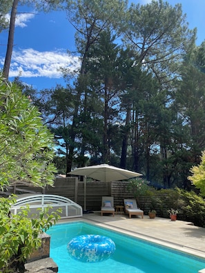 A swimming pool with a blue inflatable ring, surrounded by trees and a covered seating area with chairs and a table. - Spacious Landes villa with swimming pool, in the heart of the pines (Soorts-Hossegor)