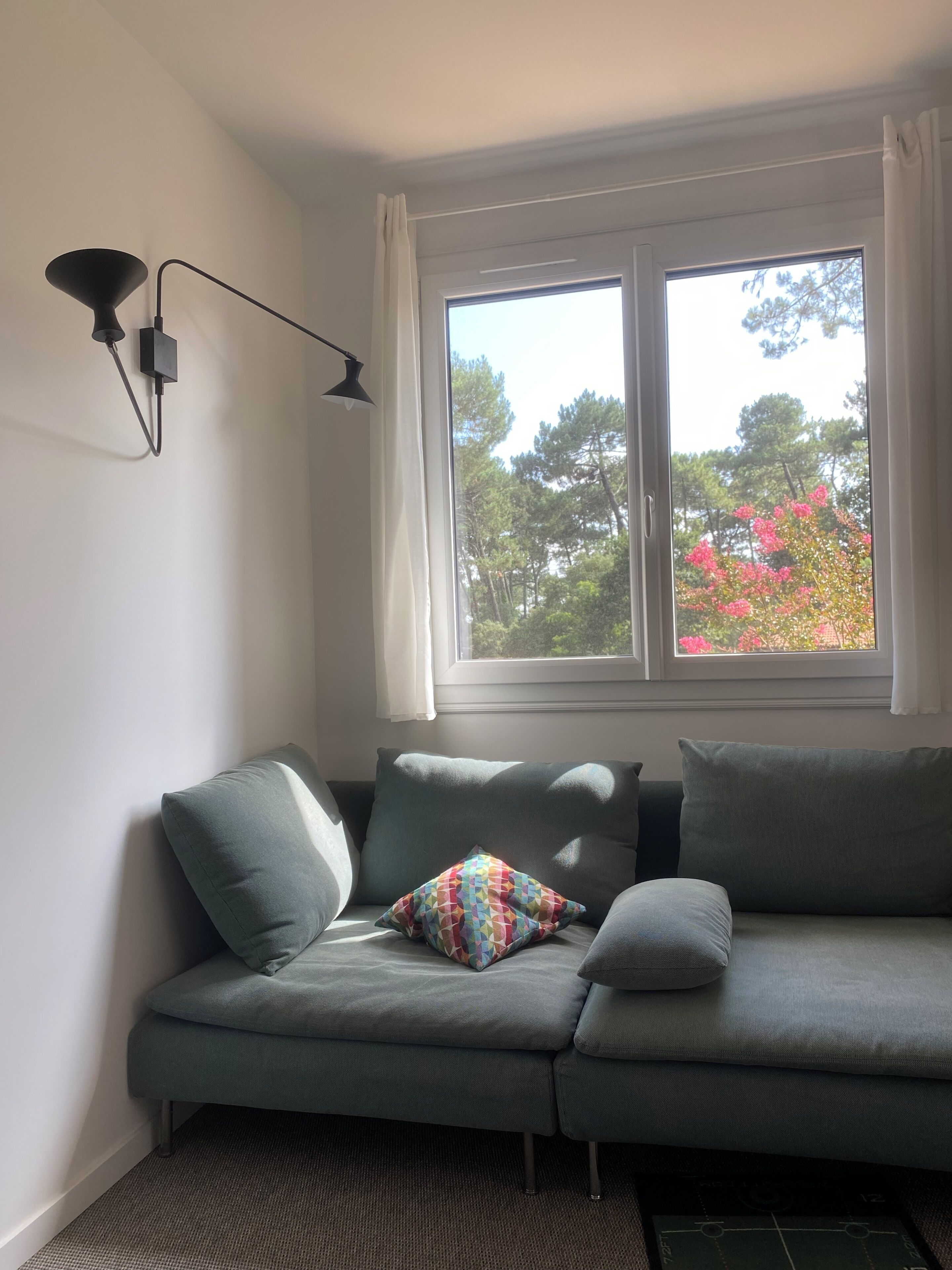 Een kamer met een grijze bank, een kussen met patroon, een zwarte vloerlamp en een raam met uitzicht op bomen en roze bloemen.