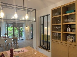 A dining area with a table set for two, a wine glass, and a cabinet with wine glasses and a coffee machine. - Spacious Landes villa with swimming pool, in the heart of the pines (Soorts-Hossegor)