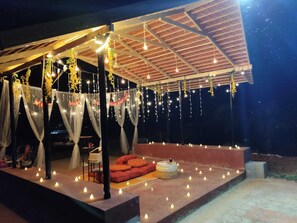 An outdoor covered area with a stage, seating, and string lights. - Anava Farms (Shrirangapattana)