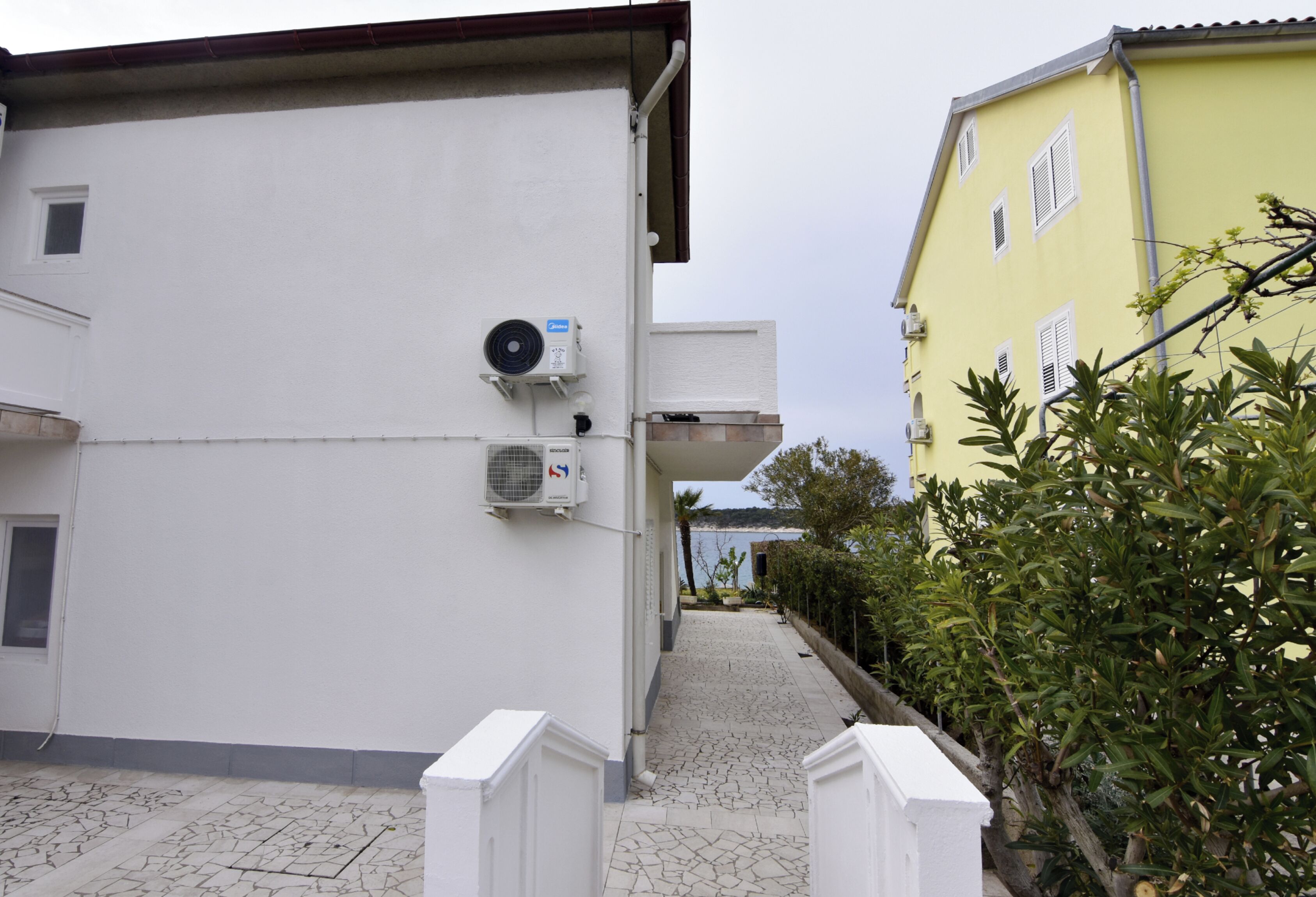 A building with a white exterior, air conditioning units, a tiled pathway, and a yellow building in the background.