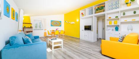 A living room with a blue sofa, a yellow sofa, a dining table with chairs, and a kitchen area with white cabinets and a yellow wall.