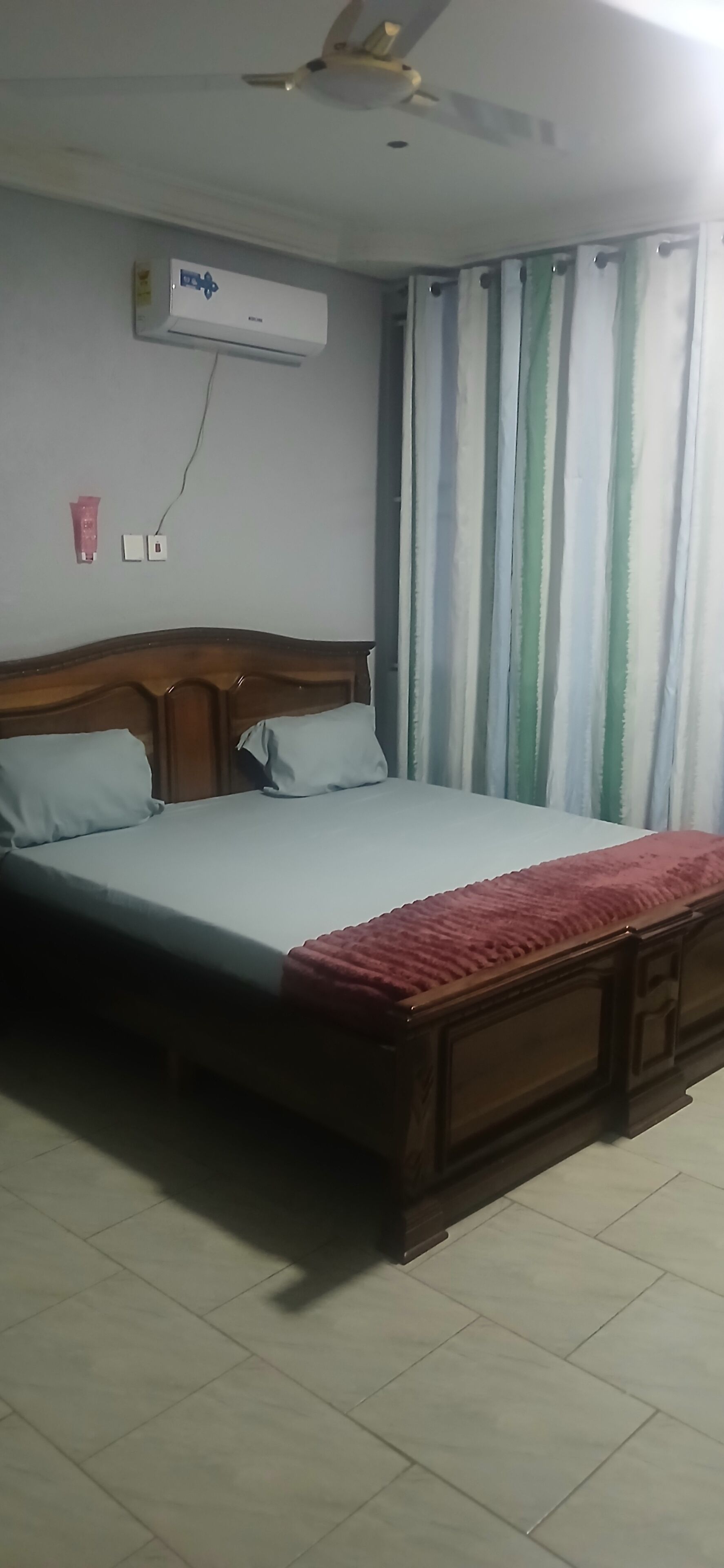 A bedroom with a bed, a ceiling fan, an air conditioning unit, and a window with curtains.