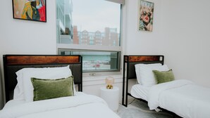 Ein Hotelzimmer mit zwei Betten, einem Fenster mit Blick auf die Stadt und einem kleinen runden Tisch mit einer Lampe.