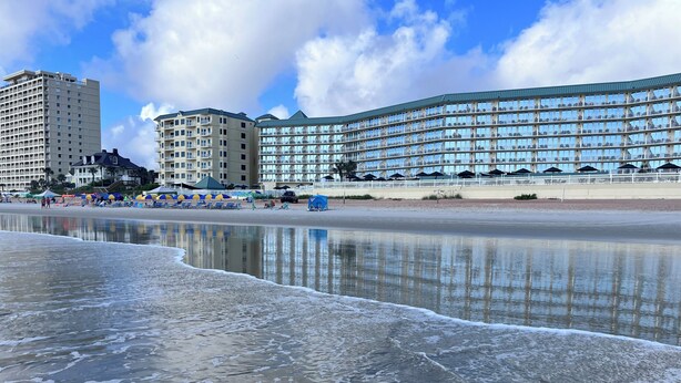 Un hôtel situé en bord de mer, doté de nombreux balcons, d’une plage de sable et d’une mer calme.