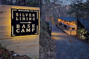 Et træskilt med teksten 'Welcome to Silver Lining Base Camp' og en pil, der peger mod en rustik hytte med metal tag.