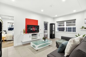 Uma sala de estar moderna com uma TV de tela plana, uma mesa de centro de vidro, um sofá cinza e um rack branco para eletrônicos.