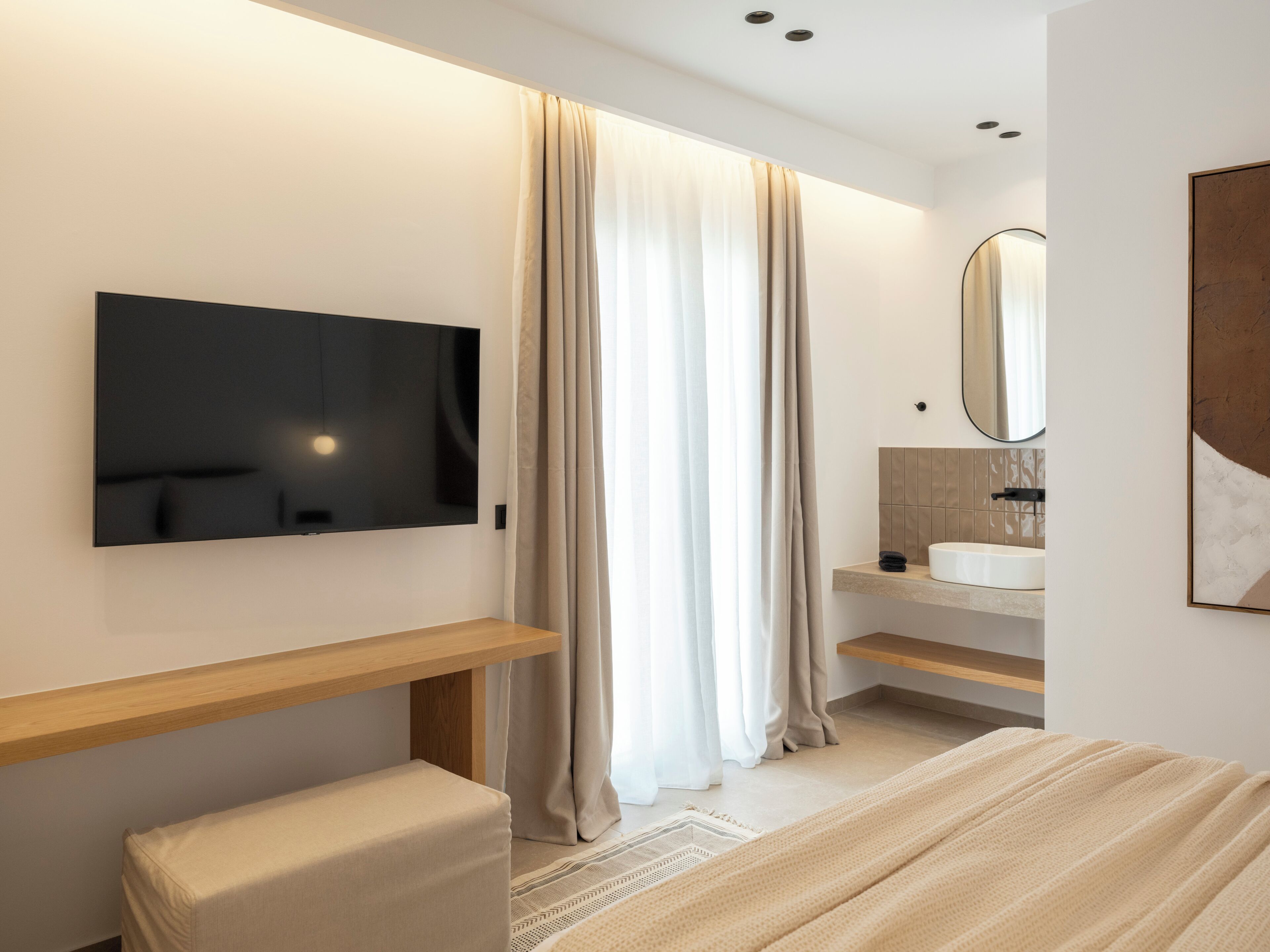 A modern bedroom with a flat-screen TV, a bedside table, a bench, a large window with curtains, a bathroom with a sink and mirror, and a framed artwork on the wall.