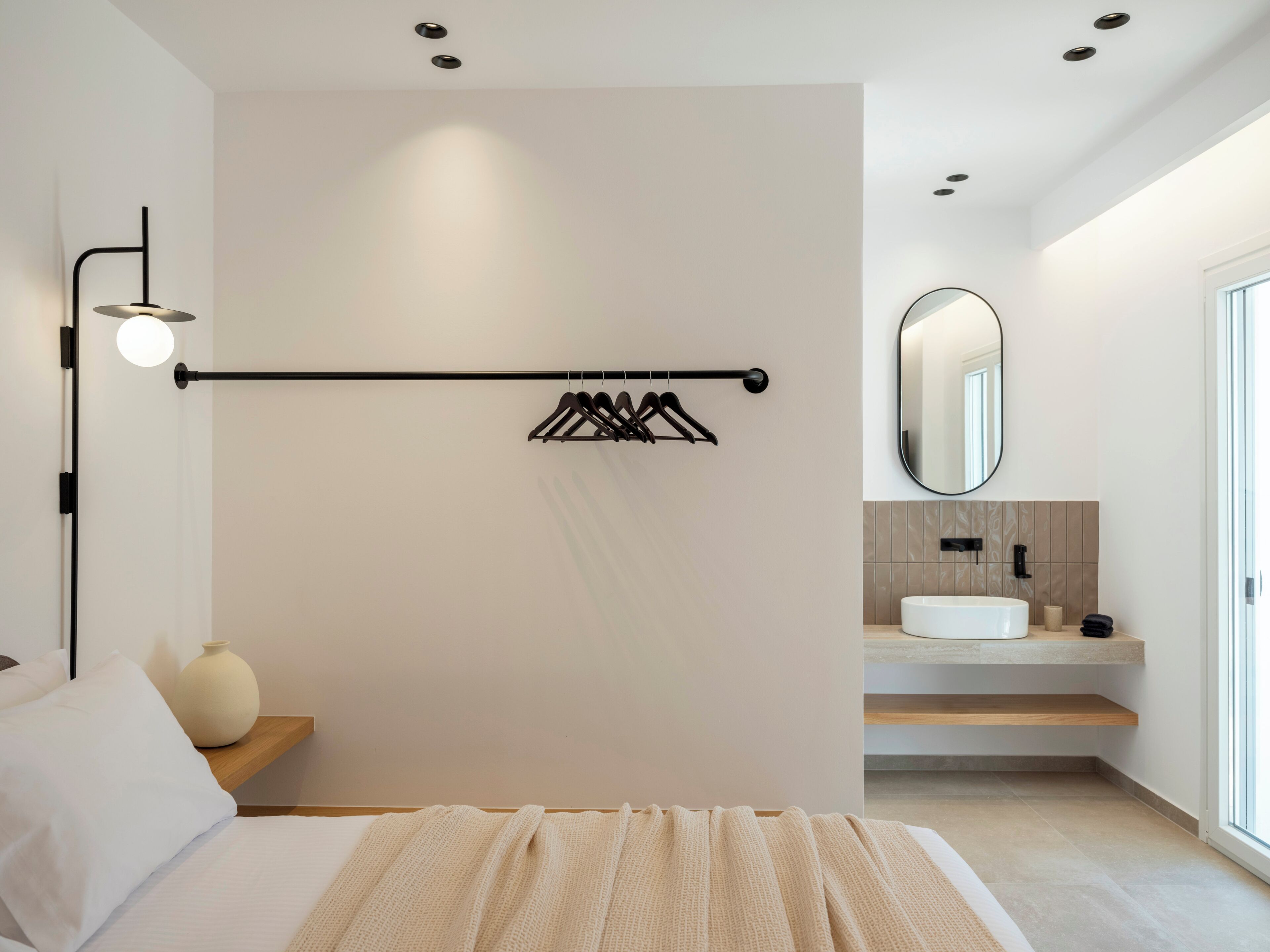 Un dormitorio moderno con cama, lámpara de noche, toallero en la pared, y un baño con lavabo y espejo.