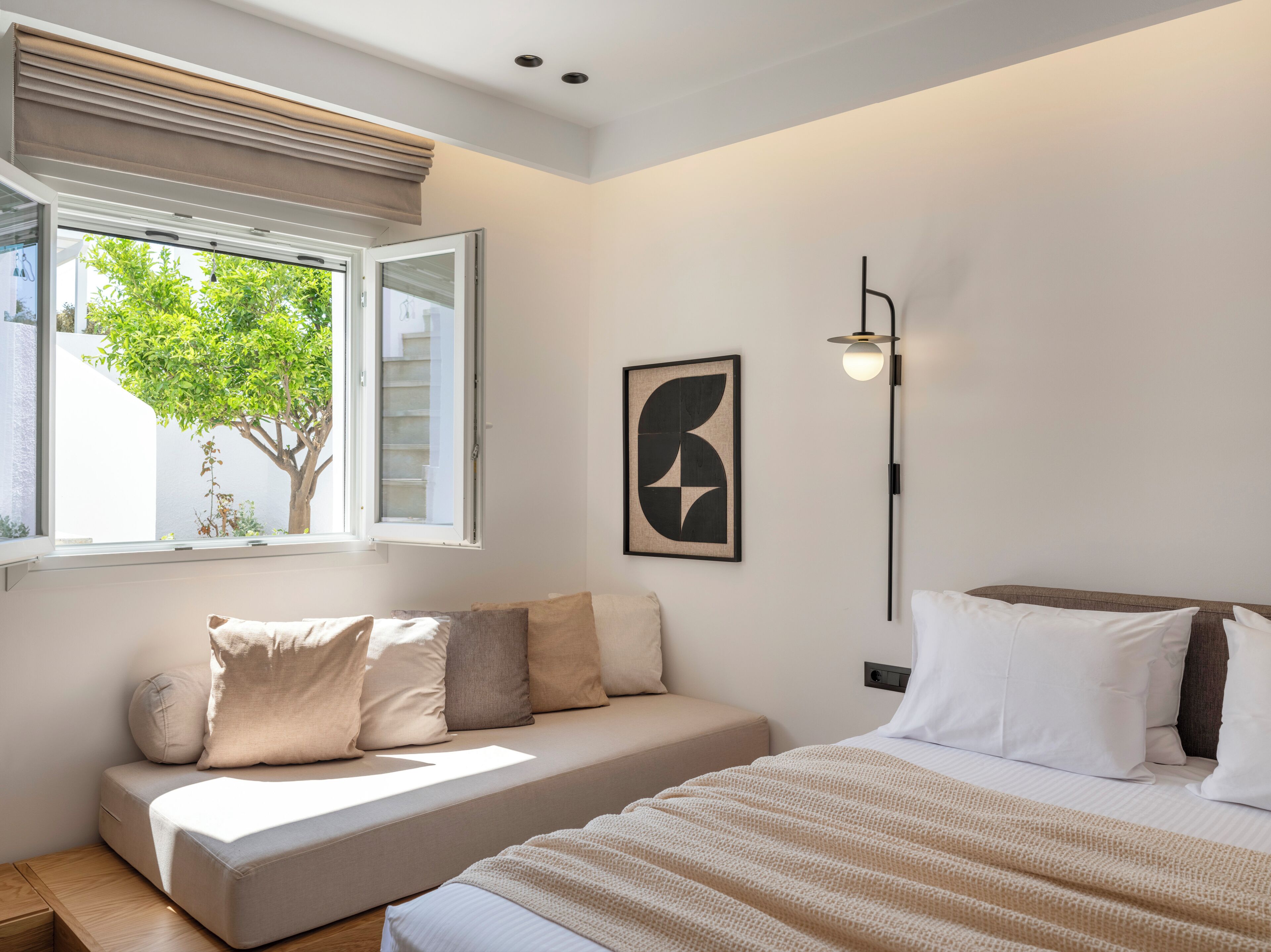 A modern bedroom with a sofa bed, a bedside table, a lamp, and a window with a view of greenery.