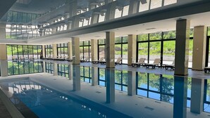 An indoor swimming pool with clear glass walls, lounge chairs, and a view of greenery outside.