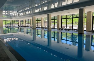 Una piscina interior con paredes de vidrio transparente, sillas de descanso y vistas a zonas verdes.