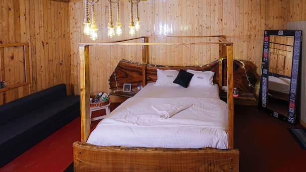 A wooden four-poster bed with white linens, a bedside table with a lamp, and a mirror on the wall. - The Buddha Village (Chikkaballapur)