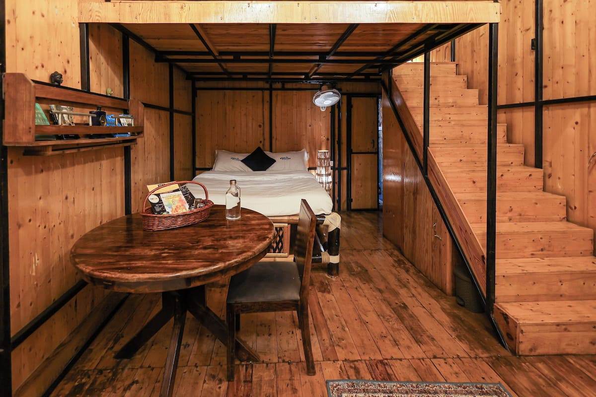 Um interior de madeira com uma cama, uma mesa com cadeiras, uma escada e uma prateleira com objetos.