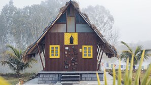 A wooden building with a triangular roof, a central door with a yellow panel, and two windows with yellow frames. - The Buddha Village (Chikkaballapur)