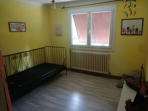 Une pièce avec un banc en métal noir, une fenêtre ornée de rideaux roses, un radiateur et un mur sur lequel est accroché un tableau.