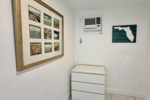 A room with a white wall, a framed collage of beach photos, a white air conditioning unit, and a white chest of drawers.