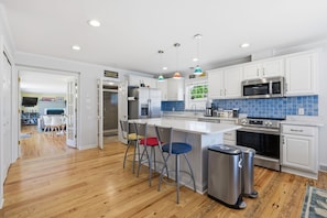A modern kitchen with white cabinets, a stainless steel refrigerator, a microwave, and a stove. There are three stools at the island, and a trash can is visible. - 3BR with POOL, Hot Tub, 2 baths, open kitchen, pkg (Rehoboth Beach)