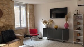 A modern living room with a stone wall, a red chair, a grey sofa, a TV, and a cabinet with decorative items. - Charming apartment in the historic center of Montignac-6 people -3 bedrooms (Montignac-Lascaux)