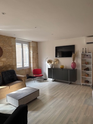A modern living room with a stone wall, a red chair, a grey sofa, a TV, and a cabinet with decorative items. - Charming apartment in the historic center of Montignac-6 people -3 bedrooms (Montignac-Lascaux)