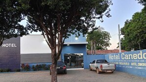 A hotel entrance with a tree in the foreground, a blue wall with the hotel's name, and a parked car. - Hotel Canedo (Corumbaíba)
