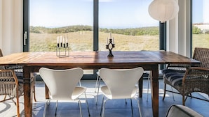 A dining area with a wooden table, white chairs, a wicker chair, and a view of the beach through large windows. - 5 Star Holiday Home in Vejby (Vejby)