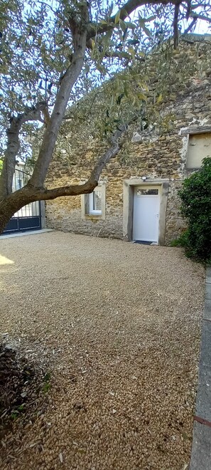 Une allée de gravier mène à un bâtiment en pierre doté d’une porte blanche, entouré d’arbres et d’un mur de pierre.