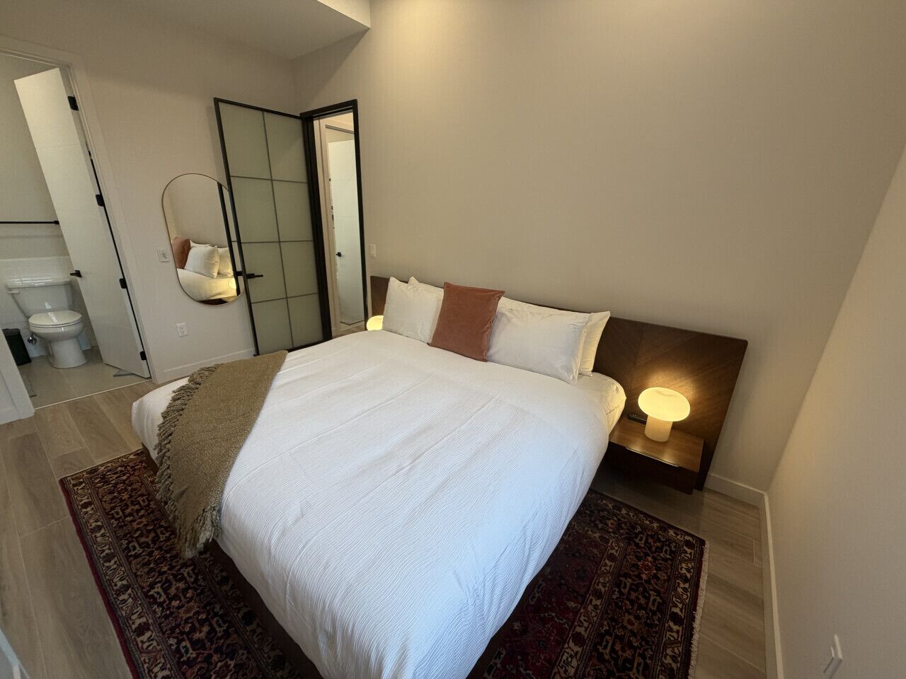 A bedroom with a bed, a bedside table with a lamp, a mirror, and a door leading to a bathroom.