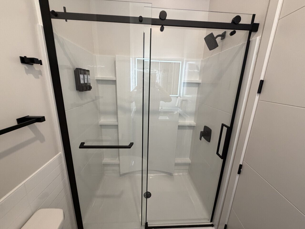 A glass-enclosed shower stall with a black frame, a white tiled wall, and a black towel rack.