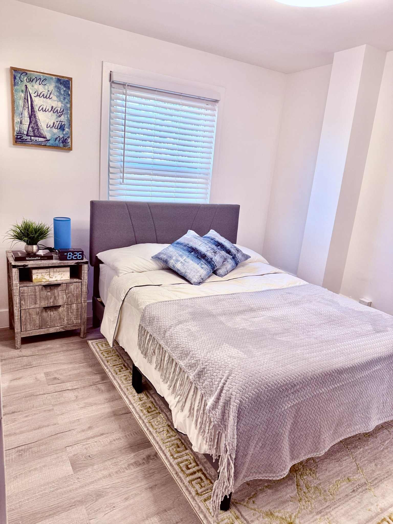 A bedroom with a bed, a nightstand with a clock, a window with blinds, and a wall art piece.
