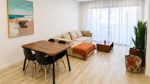 A modern living room with a wooden dining table, a beige sofa, and a large window with sheer curtains. - Modern T2 apartment with rooftop pool (Praia)