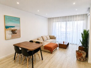 A modern living room with a wooden dining table, a beige sofa, and a large window with sheer curtains. - Modern T2 apartment with rooftop pool (Praia)