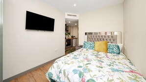 A bedroom with a bed, a flat-screen TV, a tufted headboard, and a small kitchenette in the background. - Charming Studio 1 Min Walk to Ilford Station (London)