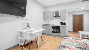 A compact hotel room with a kitchenette, a bed with a tropical print bedspread, a white dining table with chairs, and a TV mounted on the wall. - Stylish Serviced Apartment w/ Rooftop Sky Bar (London)